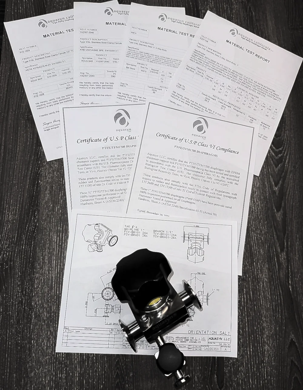 The documentation pack for one Aquasyn order laid out flat: four EN 10204 3.1 material test reports showing heat numbers and chemical analysis, two USP Class VI certificates for the PTFE/TFM1700 diaphragms, and the general arrangement drawing, with the finished valve resting on the drawing.
